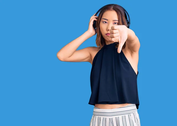 若い美しい中国の女の子は怒っている顔を持つヘッドフォンを使用して音楽を聴いて 否定的な兆候は親指で嫌いを示す 拒否の概念 — ストック写真