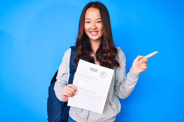 Young beautiful chinese girl showing a failed exam smiling happy pointing with hand and finger to the side