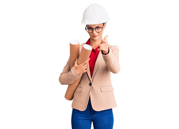 年轻美丽的女人戴着建筑师的硬礼帽 戴着眼镜 手里拿着设计图 手指着相机 对着你 自信地摆出一副严肃的姿势 — 图库照片