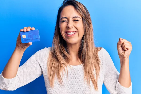 Young beautiful woman holding credit card screaming proud, celebrating victory and success very excited with raised arm