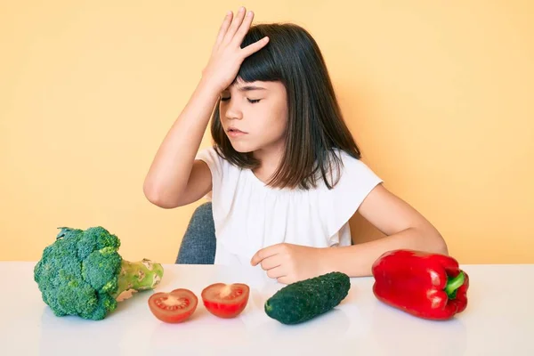 Young little girl with bang sitting on the table with veggies surprised with hand on head for mistake, remember error. forgot, bad memory concept.