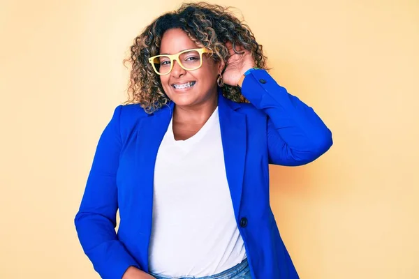 Young african american plus size woman wearing business jacket and glasses smiling with hand over ear listening and hearing to rumor or gossip. deafness concept.