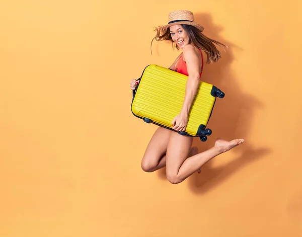 ビキニと帽子を身に着けて休暇中の若い美しい女性が幸せ 顔に笑顔でジャンプ隔離された背景にキャビンバッグを保持 — ストック写真