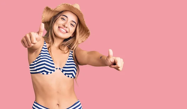 Young beautiful blonde woman wearing bikini and hat approving doing positive gesture with hand, thumbs up smiling and happy for success. winner gesture.