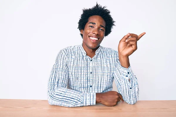 ハンサムなアフリカ系アメリカ人男性とアフロの髪を身に着けていますカジュアル服を着てテーブルの上に座って笑顔幸せなポインティングで手と指で側面 — ストック写真