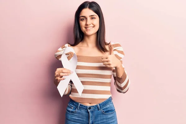 Young beautiful girl holding white cancer ribbon smiling happy and positive, thumb up doing excellent and approval sign