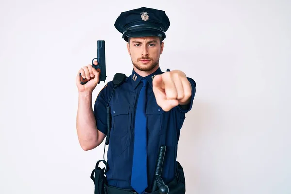 Young caucasian man wearing police uniform holding gun pointing with finger to the camera and to you, confident gesture looking serious
