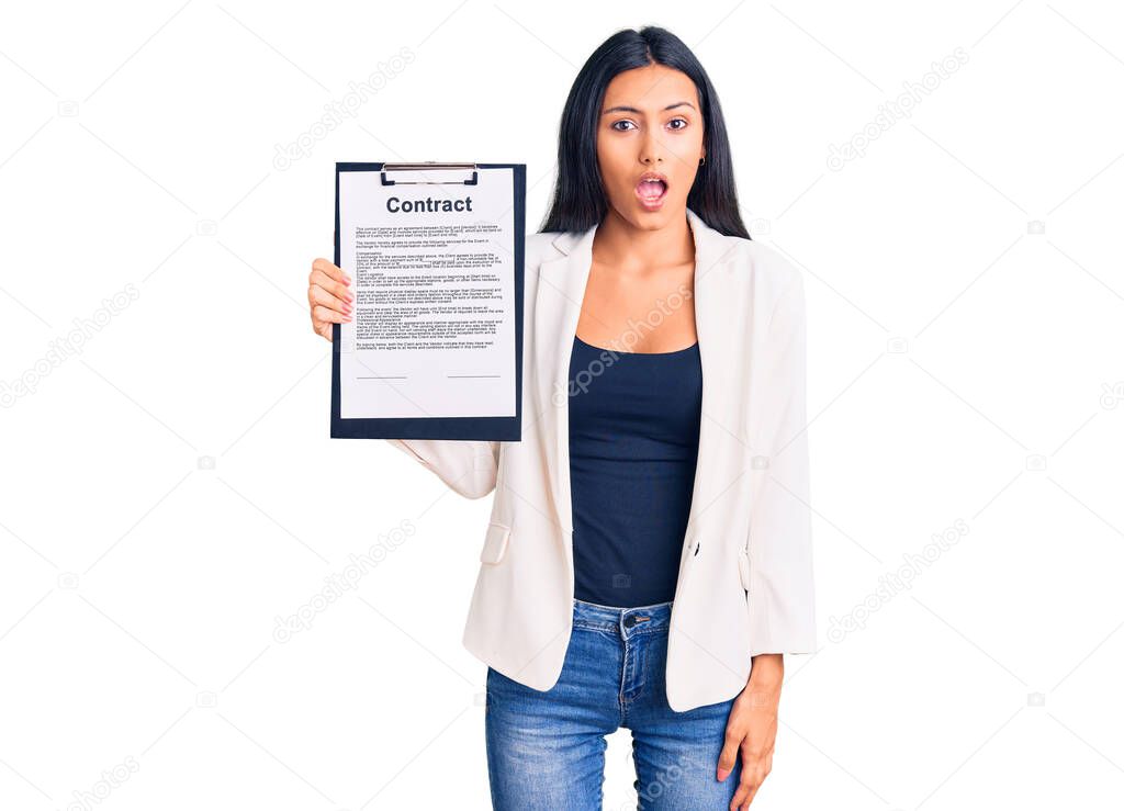 Young beautiful latin girl holding clipboard with contract document scared and amazed with open mouth for surprise, disbelief face 