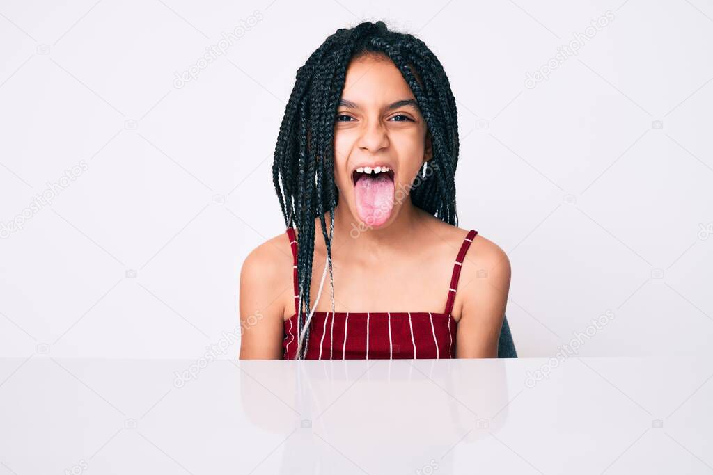 Young african american girl child with braids wearing casual clothes sitting on the table sticking tongue out happy with funny expression. emotion concept. 