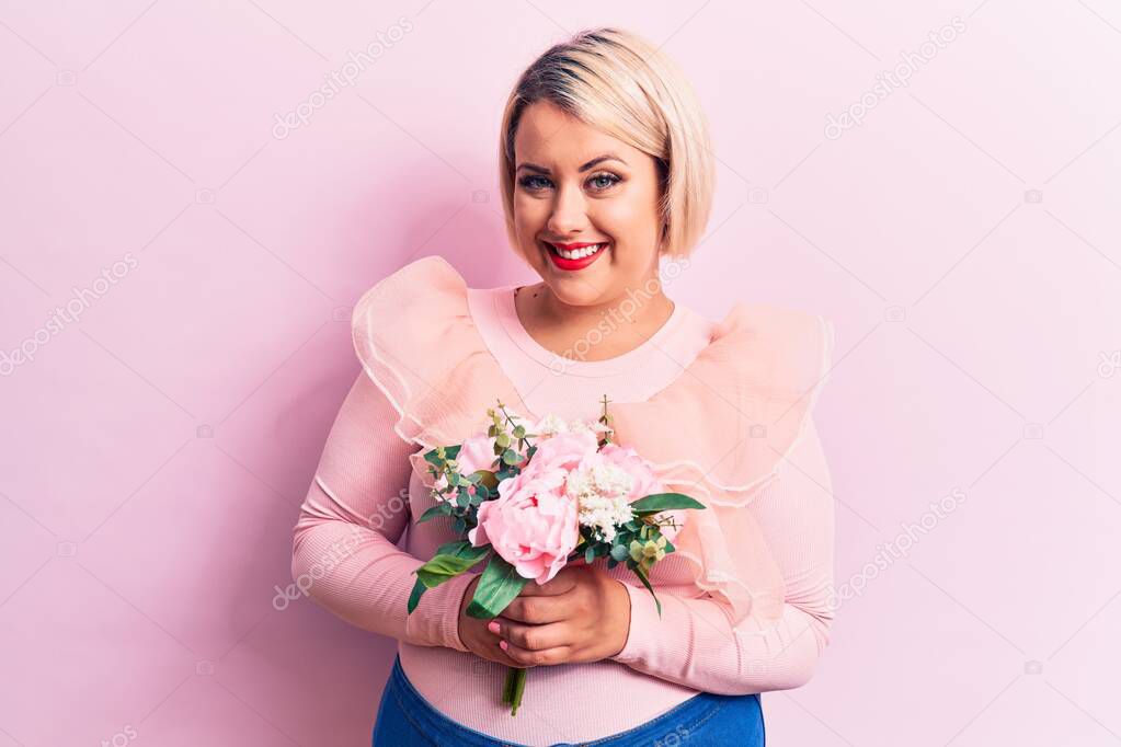 Beautiful blonde plus size woman holding bouquet of pink flowers over isolated background looking positive and happy standing and smiling with a confident smile showing teeth