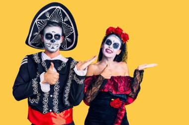 Arka planda Meksika 'daki ölü kostümünü giyen genç çift avuç içini gösteriyor ve baş parmaklarıyla hareket ediyor, mutlu ve neşeli gülümsüyor. 