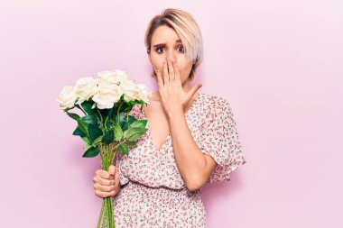 Genç, sarışın bir kadın elinde çiçeklerle ağzını eliyle kapatıyor. Şok olmuş ve hata yapmaktan korkuyor. şaşırmış ifade 