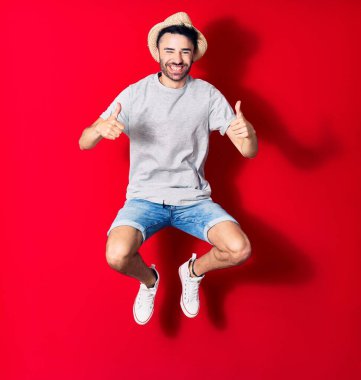 Genç, yakışıklı, günlük kıyafetler giyen ve şapkalı mutlu bir İspanyol. Yüzünde tebessümle zıplıyor. Tamam işareti yapıyor. Kırmızı arka plan üzerinde başparmaklar yukarı doğru..