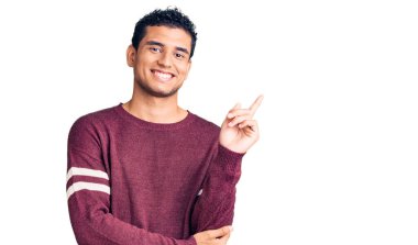 Latin asıllı yakışıklı, sıradan kıyafetler giyen, yüzünde kocaman bir gülümseme olan, elleri ve parmaklarıyla kameraya bakan genç adam.. 