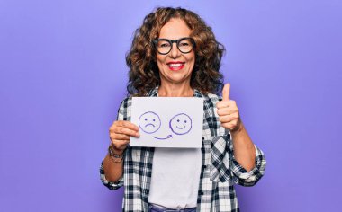 Orta yaşlı bir kadın duygu değişikliği istiyor. Elinde mutsuz, mutlu ve olumlu gülümseyen bir surat ifadesi olan bir kağıt tutuyor. Başparmağıyla mükemmel ve onay işareti yapıyor.