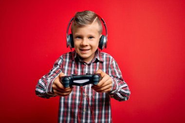Sevimli sarışın oyuncu çocuk mutlu ve kendinden emin gülümsüyor. Yüzünde tebessümle dikilip, izole edilmiş kırmızı arkaplan üzerinde gamepad ve kulaklık kullanarak video oyunu oynuyorum.