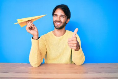 Elinde kağıt uçak tutan yakışıklı İspanyol mutlu ve pozitif gülümsüyor, başparmağıyla mükemmel ve onay işareti yapıyor. 