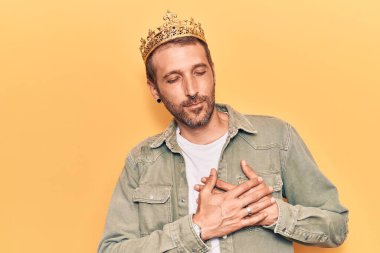 Kral tacı takan genç yakışıklı adam elleri göğsünde gülümsüyor, gözleri kapalı ve yüzünde minnet ifadesi var. sağlık kavramı. 