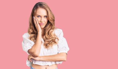 Sarı saçlı, zarif bir yaz tişörtü giyen genç beyaz kadın kollarını kavuşturup depresyondan sıkılmış ve yorgun görünüyor.. 