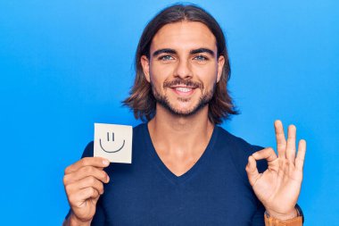Elinde gülümseme emojisi olan genç yakışıklı adam parmaklarıyla imza atıyor, gülümseyerek mükemmel bir sembol oluşturuyor. 