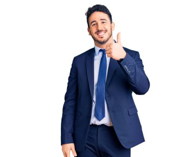 İş kıyafetleri giymiş genç İspanyol adam mutlu ve pozitif gülümsüyor, başparmağıyla mükemmel ve onay işareti yapıyor. 