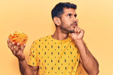 Elinde patates cipsi olan genç Latin bir adam ciddi bir yüz ifadesiyle eli çenesinde bir soru soruyor, kafa karıştırıcı bir fikir. 