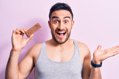 Sakallı, sakallı, enerji proteini yiyen genç bir adam izole edilmiş pembe arka planda zaferi mutlu bir gülümsemeyle ve ellerini kaldırarak kazanan ifadeyle kutluyor.