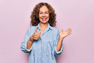 Orta yaşlı, güzel bir kadın pembe arka planda sıradan bir kot gömlek giyiyor avuç içini gösteriyor ve baş parmaklarıyla hareket ediyor, mutlu ve neşeli gülümsüyor.