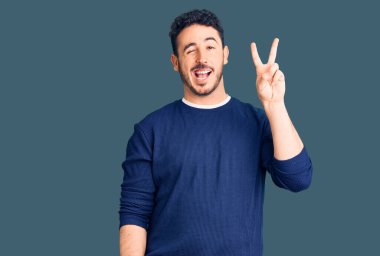 Gündelik kıyafetler giyen, gülümseyen ve kameraya göz kırpan genç İspanyol adam zafer işareti yapıyor. İki numara.. 