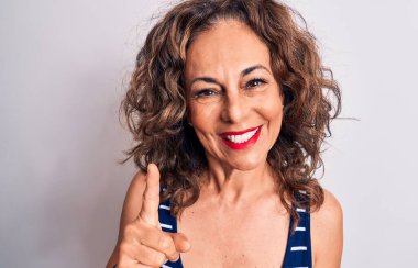 Orta yaşlı, esmer, çizgili tişört giyen güzel bir kadın beyaz arka planda duruyor. Bir fikir veya soruyla gülümsüyor ve mutlu bir yüzle parmağını kaldırıyor.