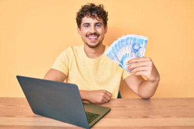 Masada oturan kıvırcık saçlı genç beyaz adam dizüstü bilgisayarla çalışıyor ve Güney Afrika banknotlarını tutuyor. Pozitif ve mutlu görünüyor ve gülümsüyor. 