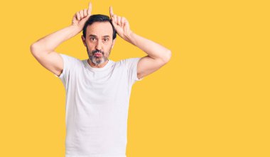 Orta yaşlı yakışıklı bir adam, sıradan tişörtler giyiyor ve parmağıyla boynuz gibi komik hareketler yapıyor. 