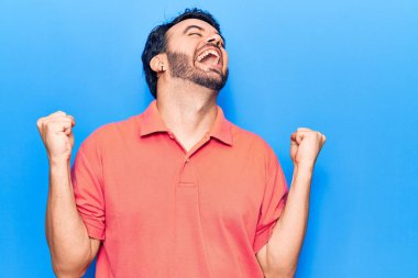 Gündelik kıyafetler giyen genç İspanyol adam çok mutlu ve heyecanlı. Kazanan jestler yapıyor. Kolları havada, gülümsüyor ve başarı için bağırıyor. kutlama konsepti. 