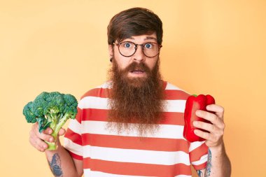 Uzun sakallı, elinde brokoli ve kırmızı biber tutan yakışıklı, genç ve kafası karışık bir adam. şüphe kavramı. 