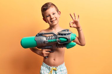 Mayo giyen sevimli sarışın çocuk su tabancası tutuyor. Tamam işareti yapıyor, gülümsüyor. Mükemmel bir sembol. 