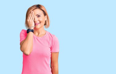 Spor giyinen genç sarışın kadın bir gözünü eliyle kapatıyor, yüzünde kendine güvenen bir gülümseme ve sürpriz bir duygu var.. 