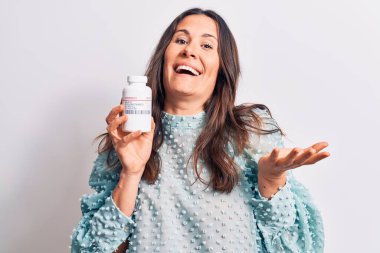 Genç, güzel, esmer bir kadın ilaç kavanozunu soyutlanmış beyaz arka planda tutarak başarıyı mutlu bir gülümsemeyle ve kazanan ifadeyle el kaldırarak kutluyor.