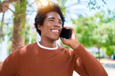 Genç, yakışıklı, Afro-Amerikan bir adam günlük giysiler giyip mutlu gülümsüyor. Yüzünde tebessümle dikilip şehrin caddesinde akıllı telefondan konuşuyor..