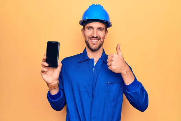 制服を着た若いハンサムな労働者の男とハード帽子を保持スマートフォンの画面を示す笑顔幸せと正 親指アップ優れた承認サインを行う — ストック写真