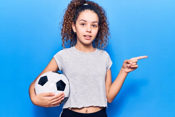 漂亮的小女孩 卷曲的头发 拿着足球开心地微笑着 手指手画脚地指向旁边 — 图库照片