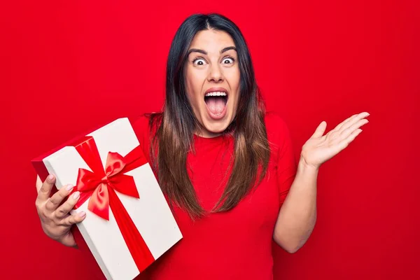 Young beautiful brunette woman holding birthday gift over isolated red background celebrating achievement with happy smile and winner expression with raised hand
