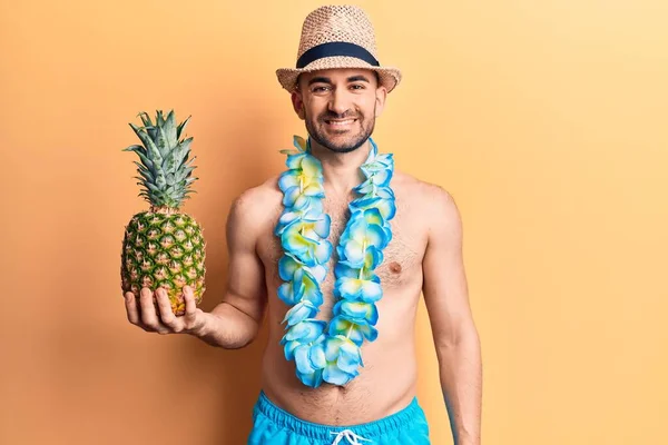 年轻英俊的秃头男子 身穿泳衣 头戴夏威夷式的莱茵 手持菠萝 站在那里 面带微笑 脸上带着自信的笑容 露出牙齿 — 图库照片