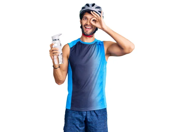 Handsome hispanic man wearing bike helmet and holding water bottle smiling happy doing ok sign with hand on eye looking through fingers