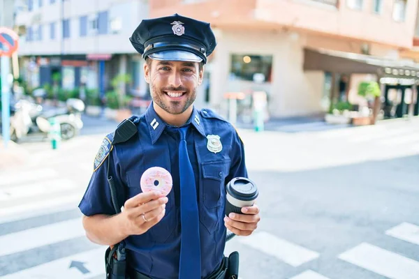 警察の制服を着た若いハンサムなパニック警官が笑顔で幸せ 町の通りでコーヒーを飲みながらドーナツを食べる — ストック写真