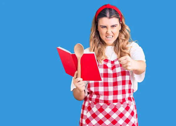 Νεαρή Καυκάσια Γυναίκα Φορώντας Ποδιά Κρατώντας Βιβλίο Συνταγών Και Κουτάλι — Φωτογραφία Αρχείου