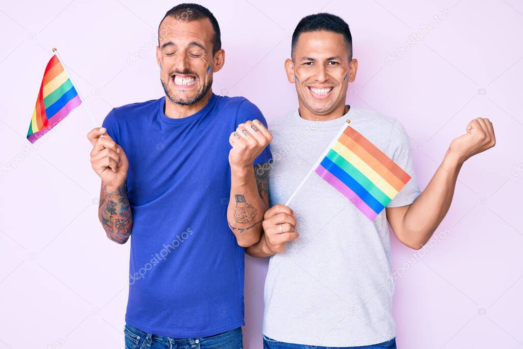 Young gay couple of two men holding rainbow lgbtq flags together screaming proud, celebrating victory and success very excited with raised arm 