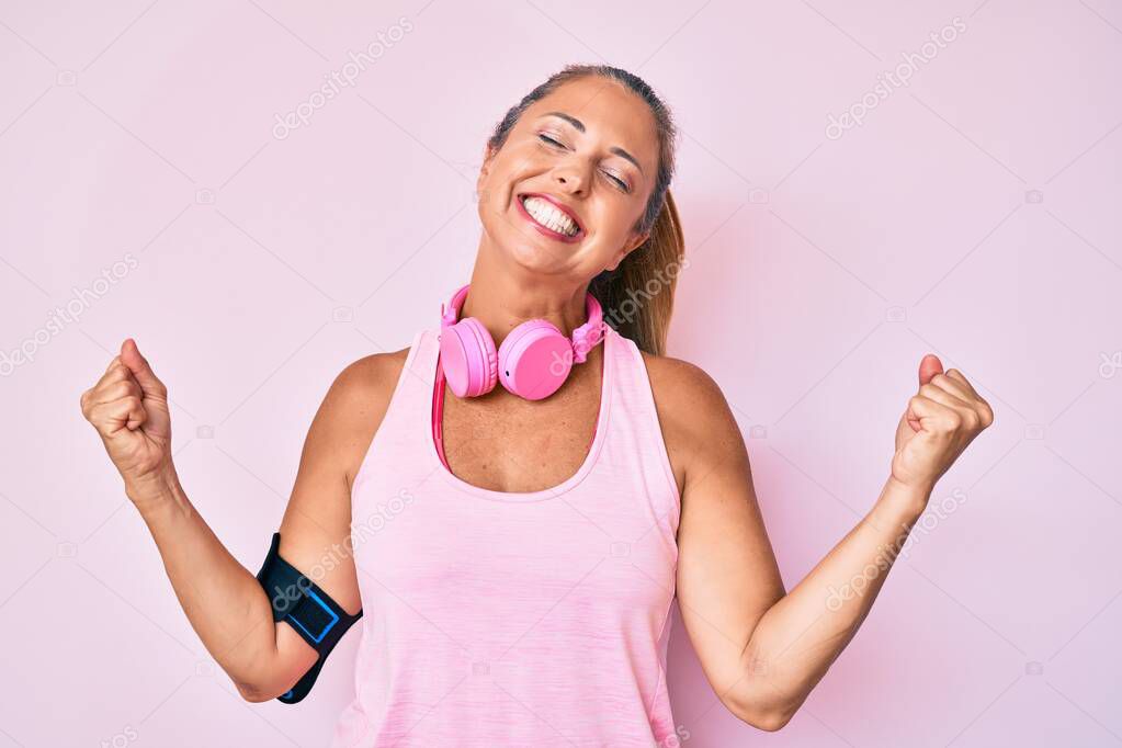 Middle age hispanic woman wearing gym clothes and using headphones very happy and excited doing winner gesture with arms raised, smiling and screaming for success. celebration concept. 
