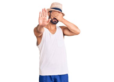 Gündelik yaz şapkası takan genç İspanyol adam elleriyle gözlerini kapatıyor ve üzgün ve korku ifadeleriyle jest yapıyor. utangaç ve olumsuz kavram. 