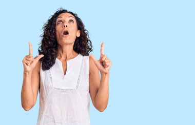 Orta yaşlı, kolsuz tişört giyen güzel bir kadın şaşkın ve şaşırmış bir şekilde yukarı bakıyor ve parmaklarıyla işaret ediyor ve kollarını kaldırıyor.. 