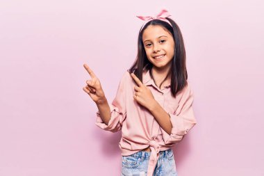 Güzel bir kız çocuğu günlük giysiler içinde gülümsüyor ve kameraya bakıyor iki eliyle ve parmaklarıyla kenarı işaret ediyor.. 